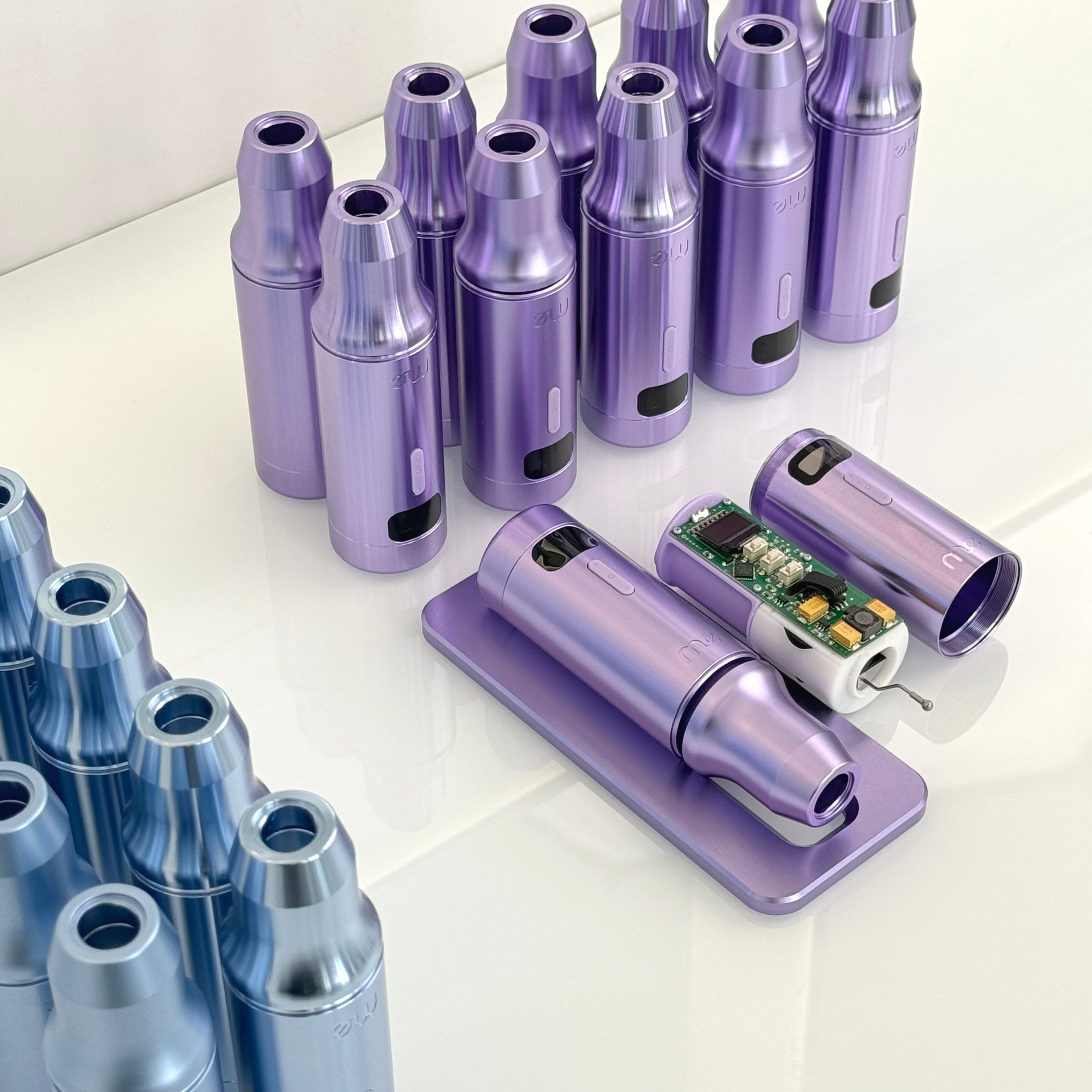 Carcasas moradas y azules de máquinas de maquillaje permanente sobre una superficie blanca, con el interior de una de ellas mostrando la electrónica.